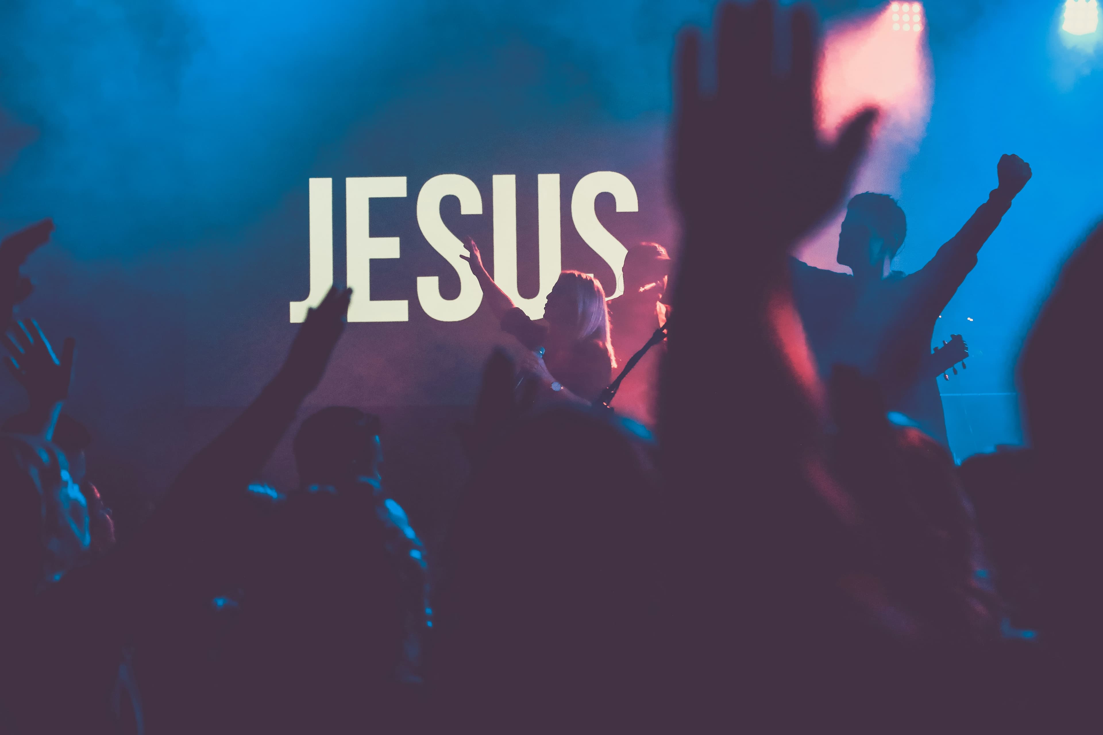 आराधना सभा में उठे हुए हाथ, और पीछे रोशनी में “JESUS” लिखा हुआ